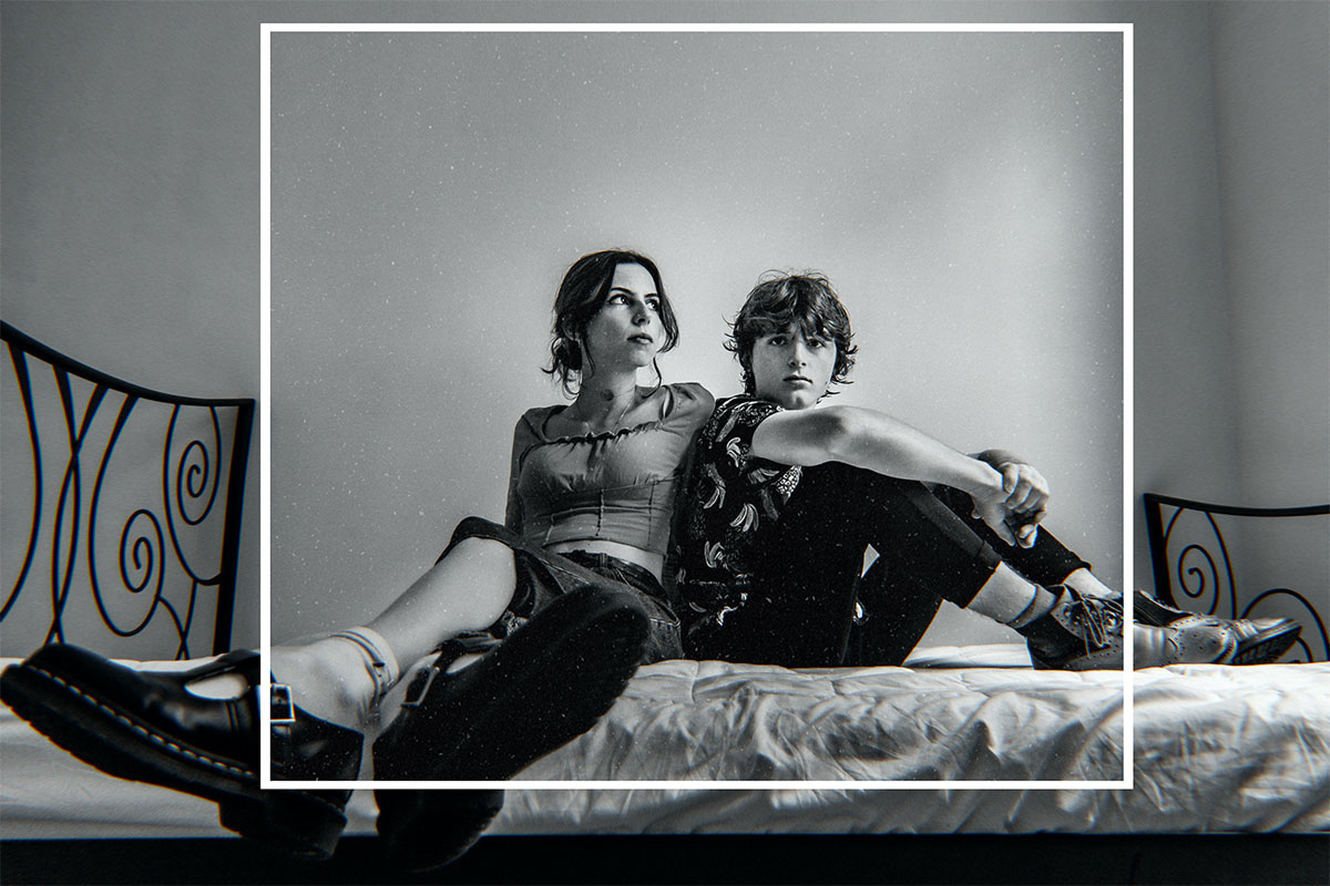 Une photo en noir et blanc d'Adèle (à gauche) et Robin (à droite) assis sur un lit. Robin a les jambes pliés, pieds chaussés sur le lit, le dos contre l'épaule d'Adèle, et regarde l'objectif. Adèle a les jambes croisées, pieds chaussés pendant du lit, et regarde vers sa gauche. Un cadre carré blanc a été ajouté numériquement par le photographe, attirant l'attention sur les sujets.