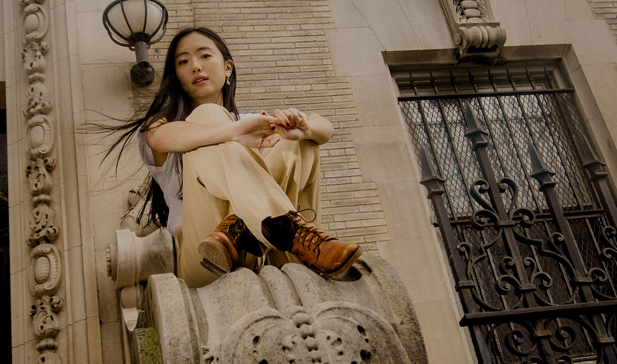 Sarah Kinsley sits, legs crossed, casually on ornate stone architecture, wearing a white t-shirt, beige pants and brown boots. A few strands of hair in the wind, she appears relaxed and introspective.