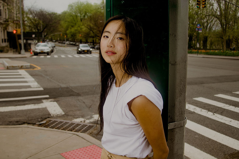 Sarah Kinsley, photographiée au coin d'une rue newyorkaise calme, appuyée contre un poteau. Vêtue d'une chemise blanche et d'un pantalon beige, elle affiche un demi-sourire qui laisse entrevoir ses dents. Elle paraît sereine.