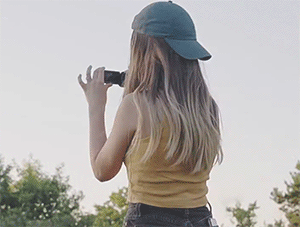 Ber turns around and bursts out laughing as she's caught filming with a camcorder. She is wearing a yellow tank top and a blue denim cap backwards.