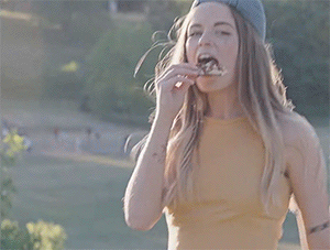 Ber dances while eating a slice of pizza outdoors. She is wearing a yellow tank top and a blue denim cap backwards. The sunlit park can be seen in the background, conveying a relaxed, casual vibe.