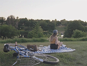 Ber is shown from the back sitting on a picnic blanket in the park, playing the ukulele. She is wearing a yellow tank top, dark denim shorts and a blue denim cap. A bicycle can be seen at the forefront while the background is all green trees and plants and shows a clear sky. The ambiance is calm and mellow.