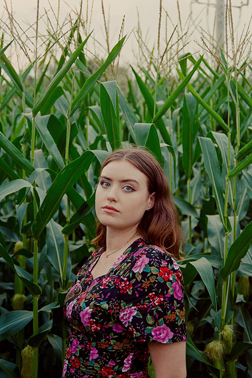 Jackie Marchal, a blue-eyed brunette, stands in a dense cornfield, wearing a floral dress. Her expression is calm and contemplative, with tall green corn stalks surrounding her.
