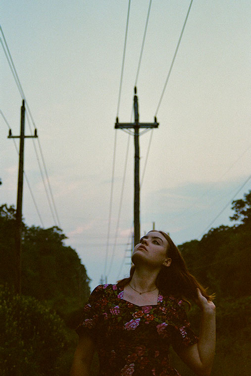 Jackie Marchal, in a floral dress, stands under power lines at dusk, gazing upward with a contemplative expression.